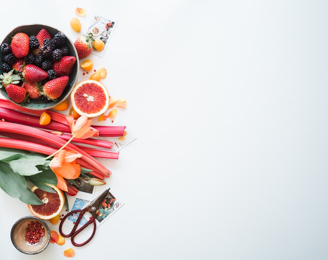 A colorful arrangement of fruits and vegetables against a white background. The assortment includes strawberries, blackberries, rhubarb, grapefruit slices, cherries, flowers, and a pair of scissors. The right side of the image has ample white space.