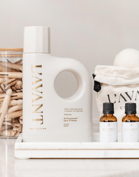 A bottle of L'AVANT high-performance laundry detergent is placed on a tray alongside two small bottles of L'AVANT products and a jar filled with clothespins. The background includes a white cloth bag labeled "L'AVANT" and a wool laundry ball.