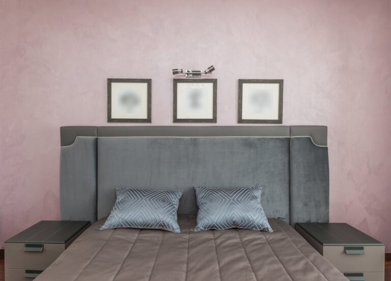 A stylish bedroom features a large gray upholstered bed with a tall headboard, two blue geometric-patterned pillows, and a gray quilt. Nightstands on each side of the bed hold lamps, and three framed pictures are arranged on the pale pink wall above the headboard.