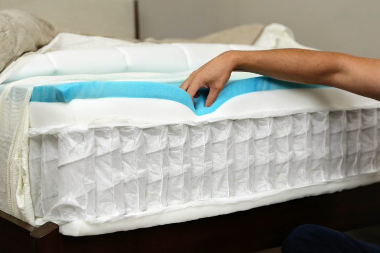 A person lifts layers of a mattress to show its construction, revealing pocket coils, a layer of white foam, and a layer of blue memory foam. The mattress is partially uncovered, exposing the various materials used for comfort and support.