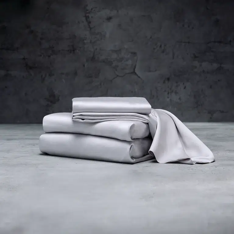 A neatly folded stack of Luxome grey bedsheets, placed on a concrete surface with a dark and slightly textured grey wall in the background. The set includes a fitted sheet, a flat sheet, and a pillowcase.