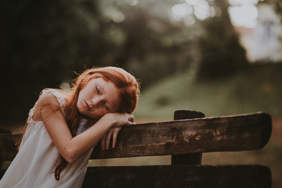 A young girl with long red hair is peacefully resting her head on her folded arms atop a wooden bench. She is wearing a white sleeveless dress, and the background shows a blurred outdoor setting with greenery, suggesting a serene, late afternoon or early evening scene.