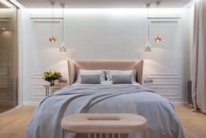 Modern bedroom with a neatly made bed featuring a light gray comforter and pillows. The bed has a beige upholstered headboard. Two pendant lights hang from the ceiling on either side of the bed. A small side table with a flower arrangement is on the left. The walls are white.