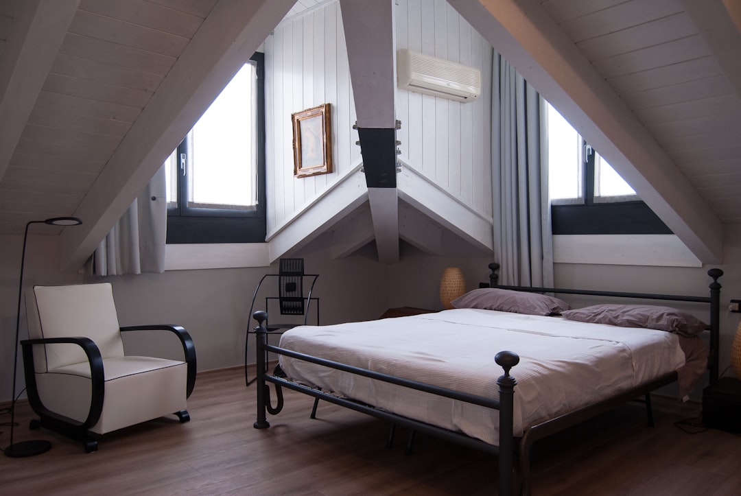 An attic bedroom with slanted ceilings, featuring a neatly made bed with a black metal frame, two windows, a white armchair with black accents, a floor lamp, and a small shelving unit. The space is accented with white and light gray tones.