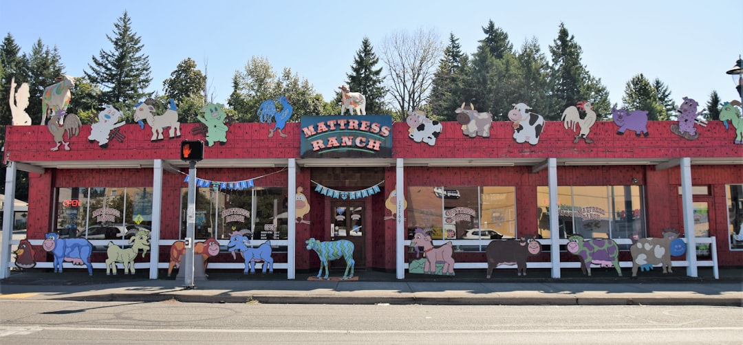 The image shows the exterior of Mattress Ranch, a store with colorful and whimsical animal cutouts decorating the facade. The animals, including cows, sheep, and horses, are painted in various cartoonish styles. Trees and a clear sky are in the background.