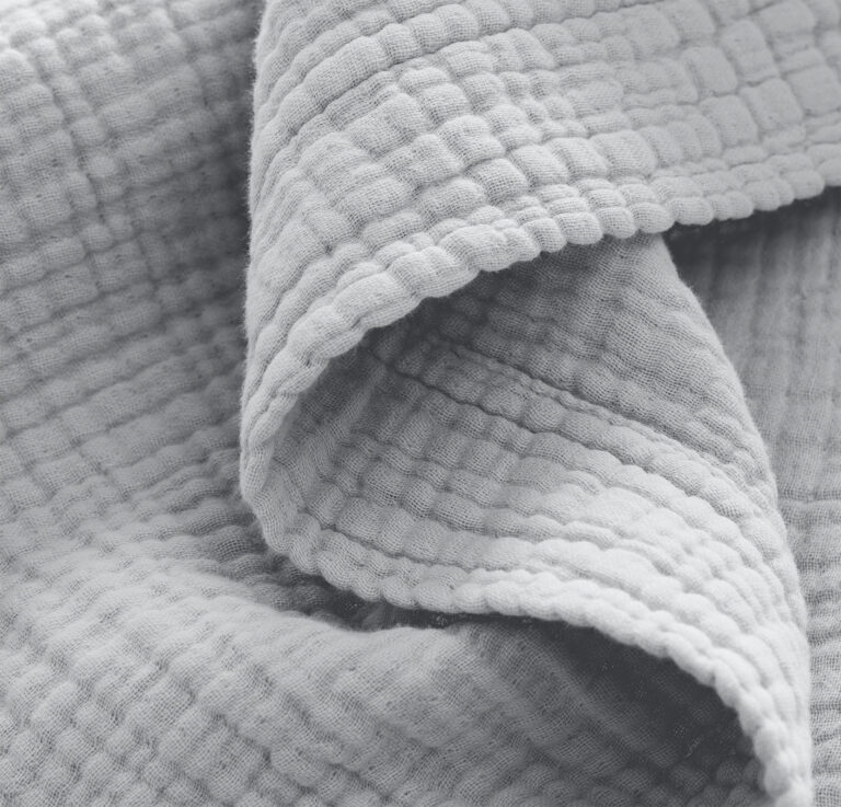 Close-up of a soft, textured fabric with a waffle weave pattern. The fabric appears crumpled and has a light, muted color. The textured details and folds of the material are prominently visible, highlighting its tactile quality.