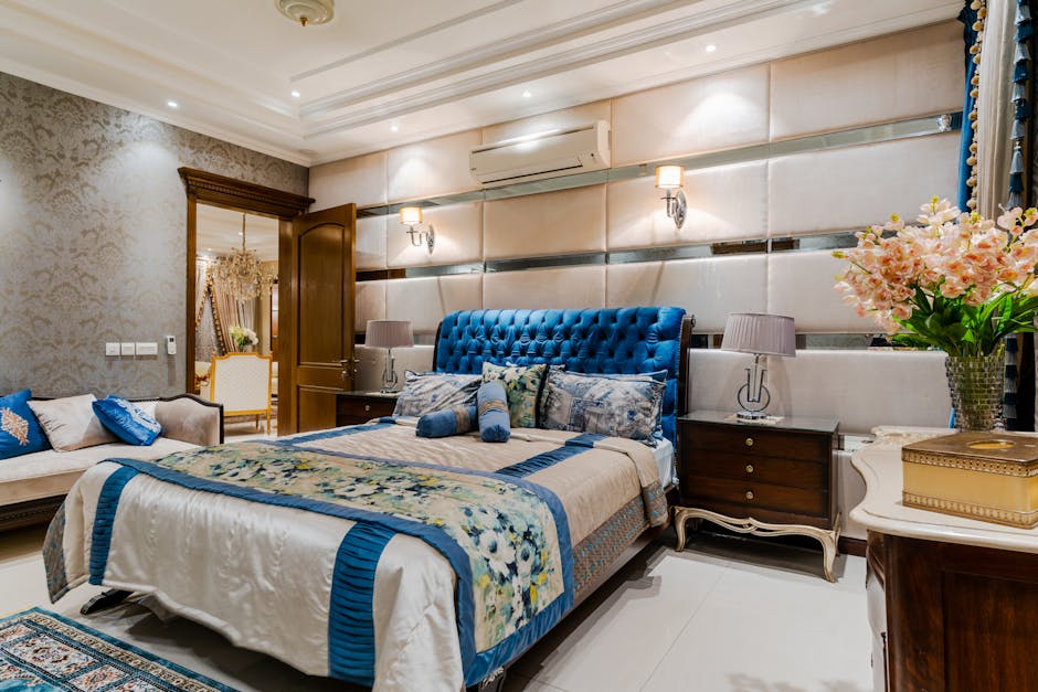 A luxurious bedroom featuring a bed with a tufted blue headboard and floral bedding. There are two bedside tables with lamps, a sofa, and a vase with flowers. The room is well-lit, with ornate wall details and a view into an adjacent room through an open door.