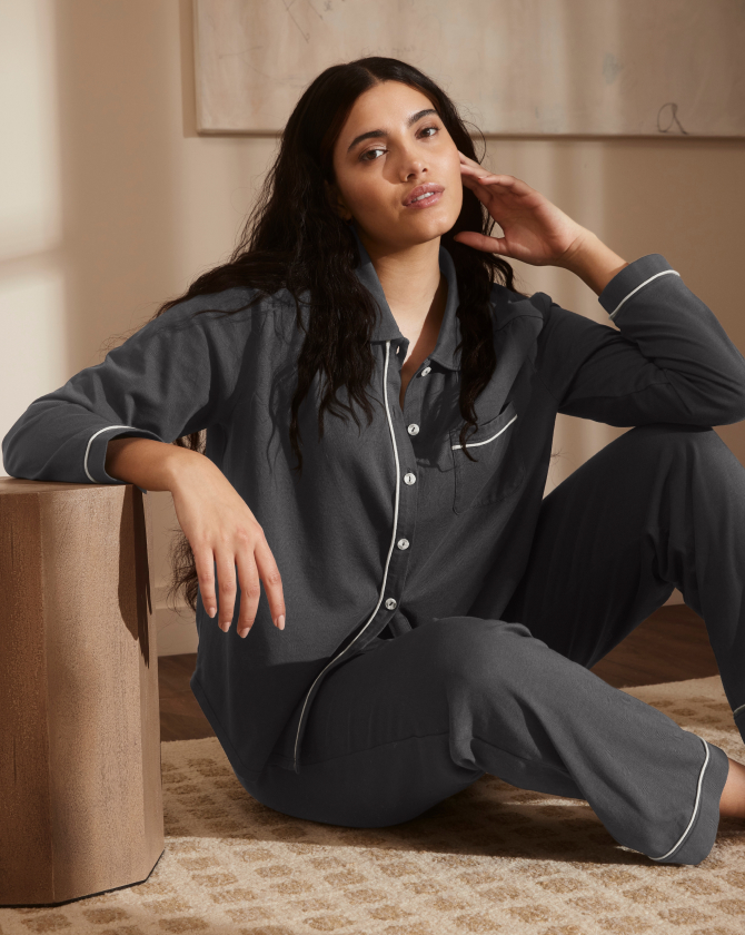 A person with long dark hair is sitting on the floor and leaning against a beige piece of furniture. They are wearing dark-colored, button-up pajamas with light trim. They have a relaxed expression and one hand rests gently on their neck.