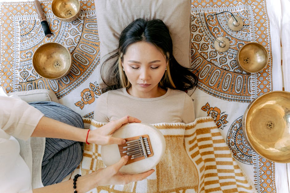 A person is lying on a patterned blanket with their eyes closed, receiving a sound therapy session. Several brass singing bowls and chimes are placed around them. Another person’s hands are positioned near the receiver, holding a small musical instrument.