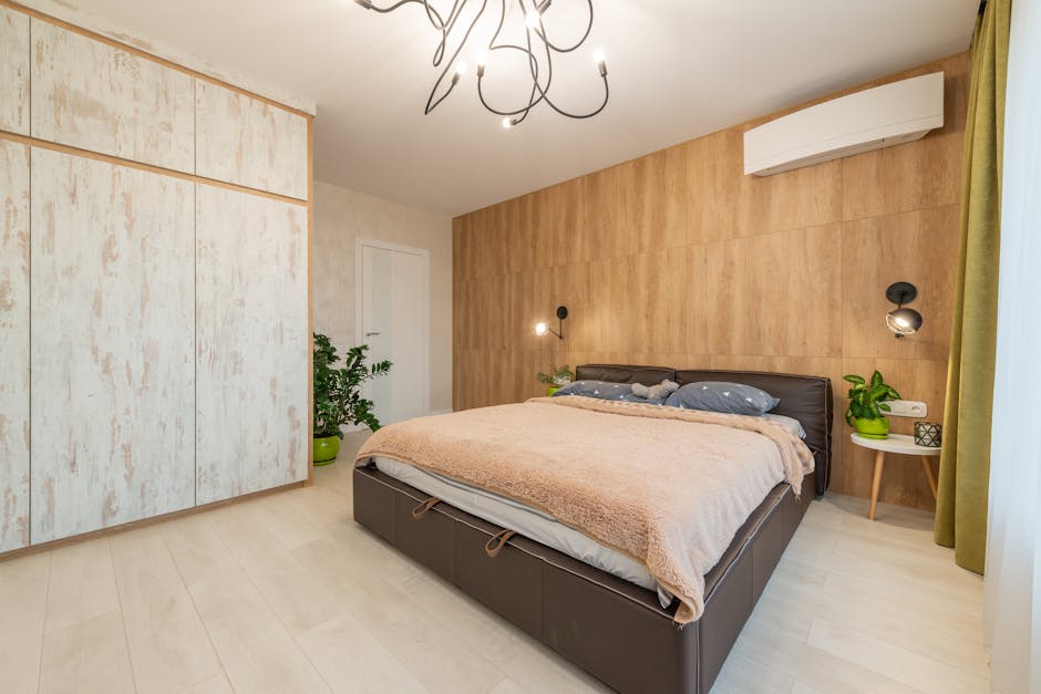 A modern bedroom with light wood walls and white wood-paneled closets. The room features a large bed with a brown leather frame and beige blanket, accented by gray pillows. Houseplants are placed nearby, with a round side table holding a plant and decorative items.