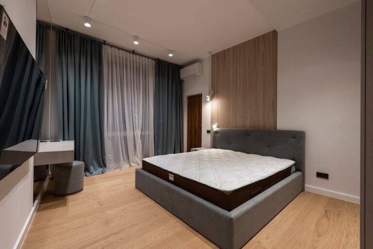 A modern bedroom with a muted color palette featuring a large bed with a gray upholstered headboard, light wood flooring, a wall-mounted flat-screen TV, a small desk with a stool, and floor-to-ceiling curtains covering a window.