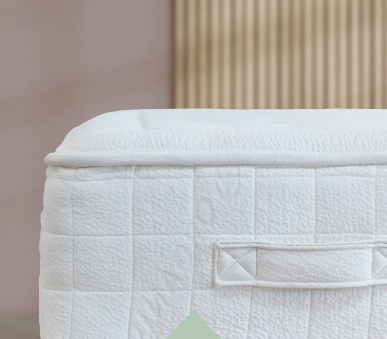 A close-up of the corner of a white, quilted mattress with a textured pattern. The mattress features a fabric handle stitched to the side. The background includes a blurred vertical wooden slat wall, providing a neutral setting.