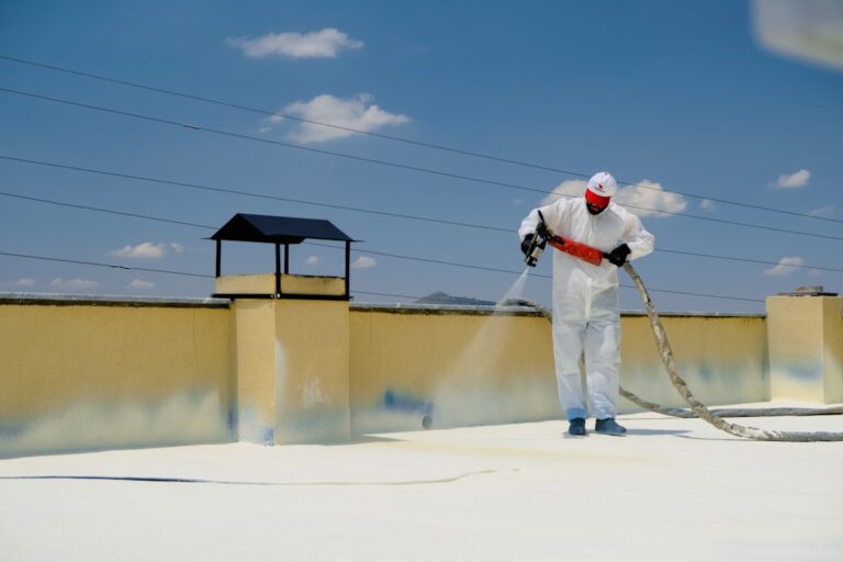 A person wearing a white protective suit and red mask sprays a roof with white foam insulation under a bright blue sky. Electrical wires run above, and there is a yellow chimney-like structure nearby. The setting appears to be on top of a building.