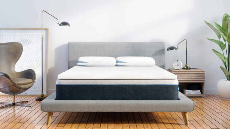 A modern, minimalist bedroom with a grey upholstered bed frame and headboard. The bed has two white pillows and a white mattress. There are sleek black wall-mounted reading lamps on both sides, a wooden nightstand with an alarm clock, and a potted plant nearby.