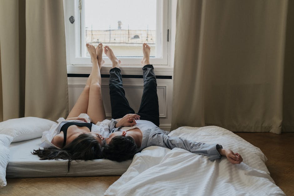 A couple lies on their backs on a mattress near a large window with their legs resting up against the window frame. They appear relaxed and are holding hands, surrounded by pillows and bedding. Sunlight softly illuminates the room.