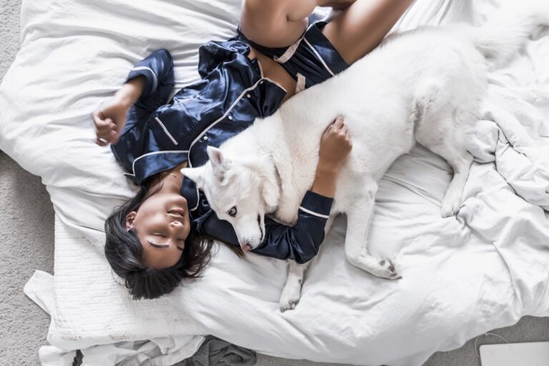 A person wearing dark blue pajamas is lying on a bed while embracing a large, white dog. The person has dark hair and appears relaxed, with one arm wrapped around the dog. The dog looks content and is lying close to the person on the white bedding.