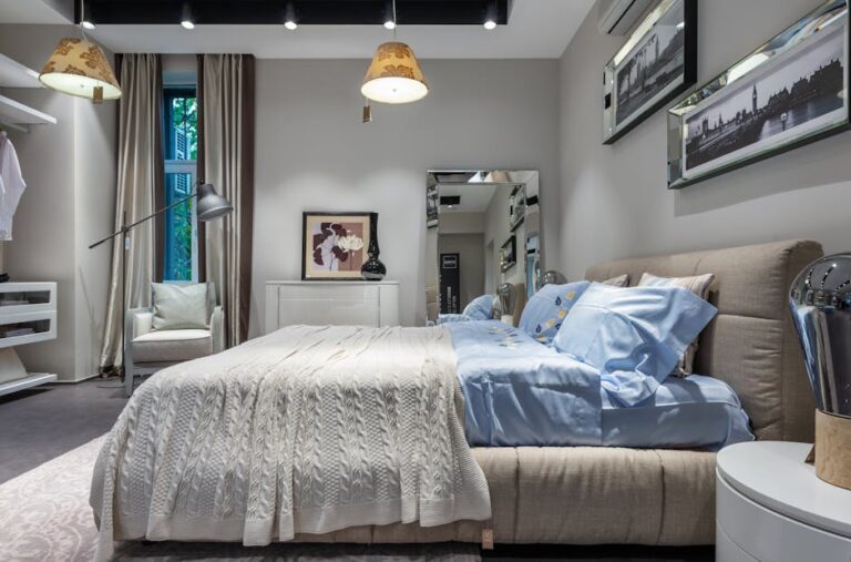 A cozy bedroom with a beige upholstered bed adorned with blue and white linens. The room features a large window with beige curtains, modern lighting, framed artwork, a standing mirror, and a white armchair. The neutral color palette creates a serene atmosphere.