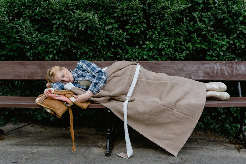 An elderly person with glasses, wearing a plaid shirt and pants, sleeps on a wooden bench covered with a blanket. The bench is in a park with a dense green hedge in the background.