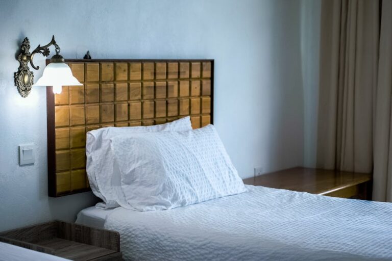 A neatly made bed with white linens stands against a light blue wall. A wooden headboard with a square pattern decorates the bed, with a vintage wall-mounted lamp providing a soft glow. A wooden table is visible in the corner, partially hidden by a beige curtain.