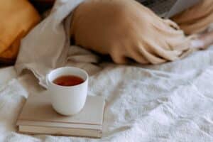 A white mug filled with tea rests on two closed books on a bed with white linens. A person, partially covered in a beige blanket and holding a book or newspaper, is visible in the background.