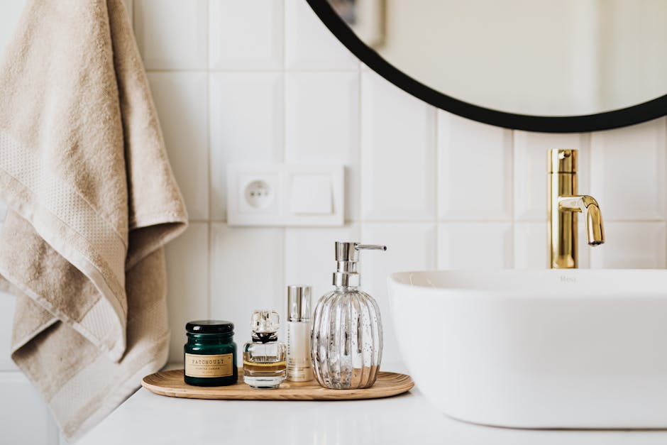 A neatly organized bathroom counter featuring a round mirror, a white sink with a gold faucet, and a wooden tray holding various toiletries including a green jar, a glass bottle, a clear spray bottle, and a clear pump bottle. A beige towel hangs nearby.