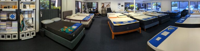 A panoramic view of a mattress store showcasing various beds on display with neatly arranged pillows and bedding. Shelves with pillows and other sleeping accessories are visible on the left, and the store has large windows letting in natural light.