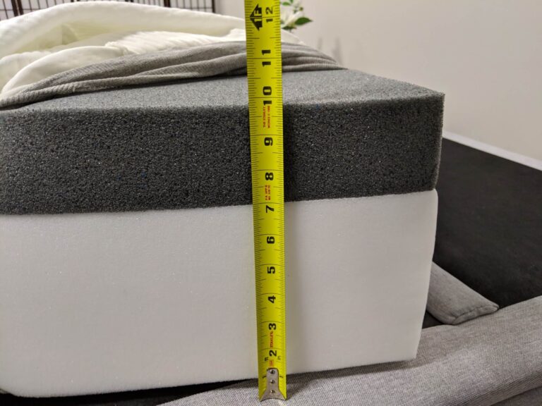 A close-up view of a layered mattress, comprised of a gray top layer and a white bottom layer. A yellow measuring tape, standing vertically alongside the mattress, shows the height measurement to be approximately 10 inches. A folded white blanket is visible on the mattress.