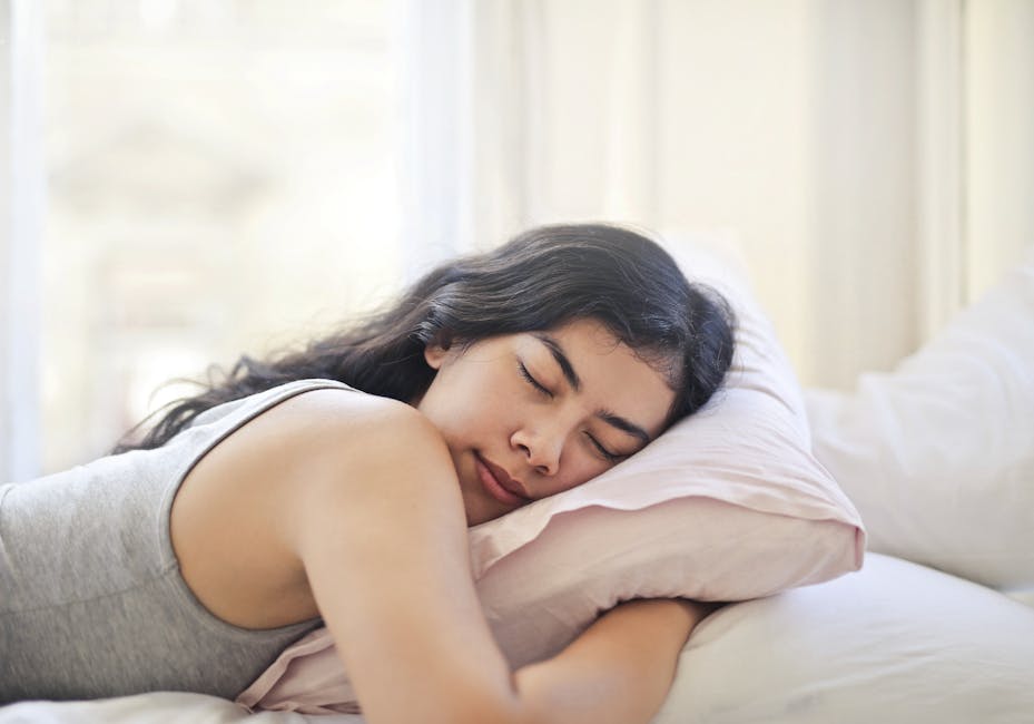 A woman with long dark hair sleeps peacefully on her side, resting her head on a pink pillow. She is wearing a gray tank top, and the background shows a bright, softly lit room with white bedding and curtains.