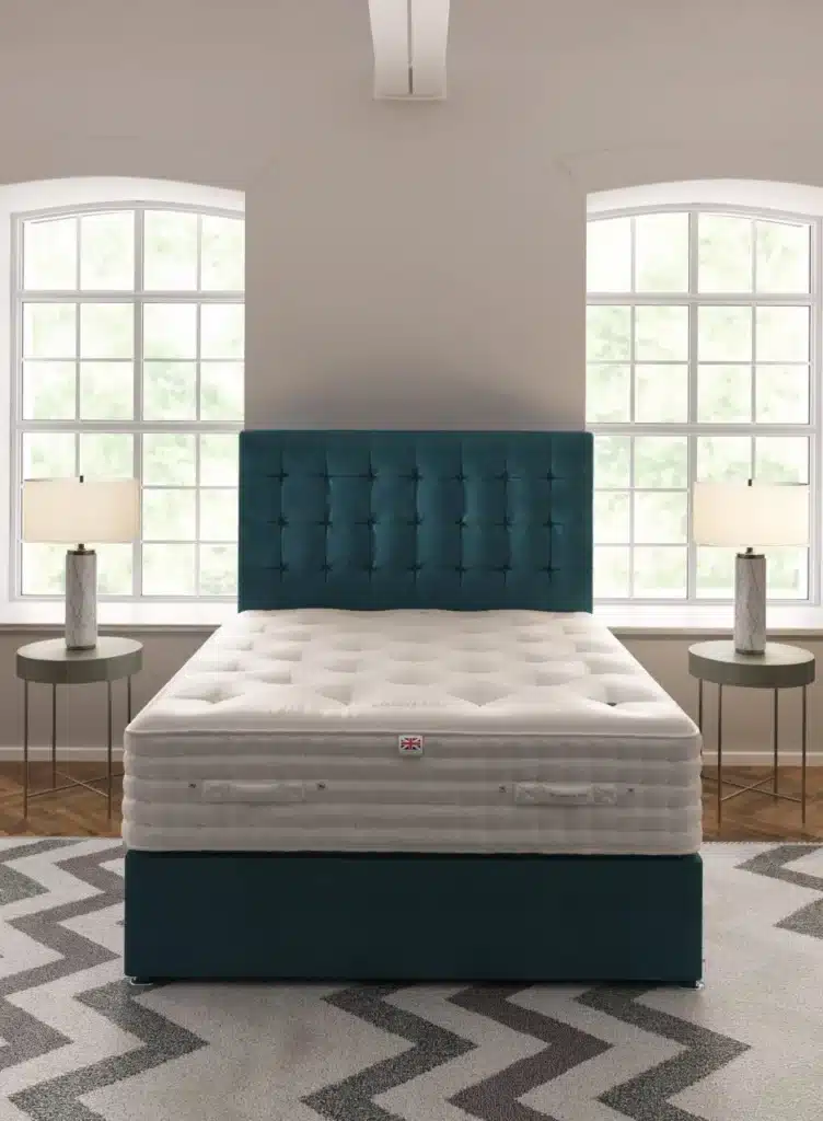 A neatly made bed with a blue tufted headboard is centered between two large windows. Each window has a table with a lamp beside it. The floor features a zigzag patterned rug and the room is decorated in neutral tones with a touch of blue from the bed.