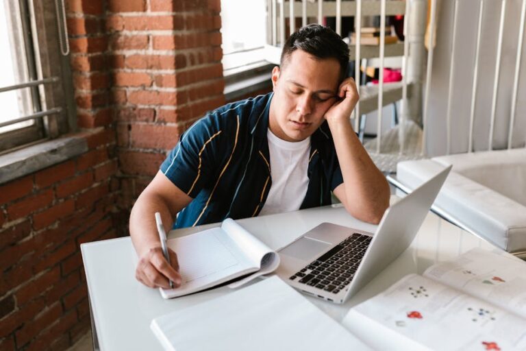 A person with short hair, wearing a striped shirt and white T-shirt, rests their head on one hand while writing in a notebook with the other hand. They sit at a white table with an open laptop and papers in front of them, next to a brick wall and window.