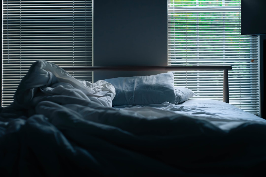 A dimly lit bedroom with an unmade bed in the center. Light streams in through partially open window blinds, casting shadows and a soft glow on the rumpled white sheets and pillows. The overall mood is serene and quiet.