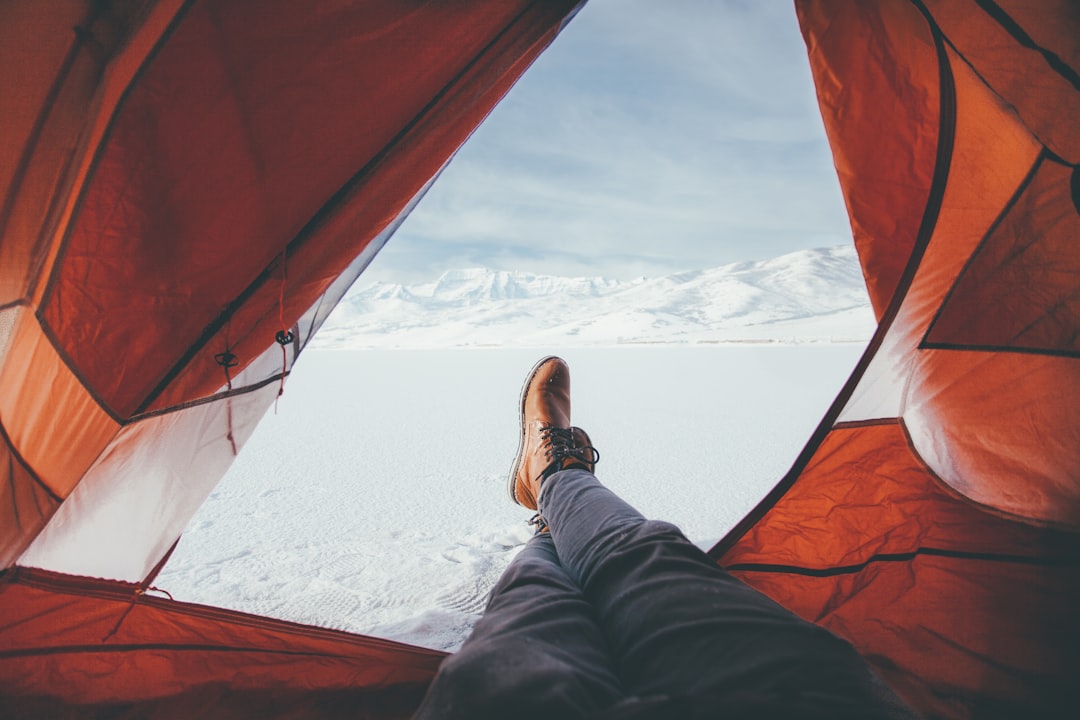 A person is lying inside a tent with their legs extended, wearing brown boots and dark jeans. The tent is open, revealing a vast snowy landscape and distant mountains under a clear blue sky.