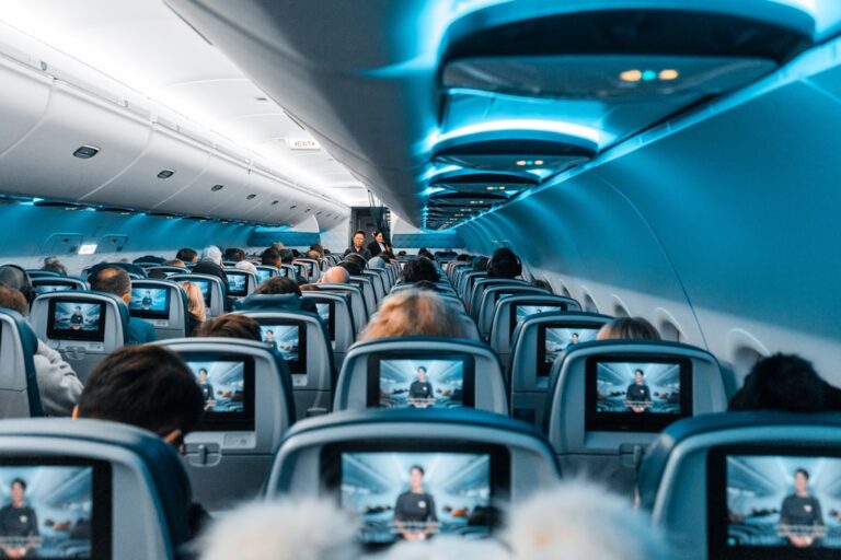 Interior of an airplane cabin filled with passengers. Each seat has an individual screen showing the same movie. The cabin is lit with blue overhead lights, giving a modern and calm ambiance. Flight attendants are seen at the far end of the aisle.