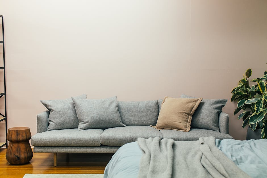 A modern living room with a light gray sofa, decorated with several cushions, one being a different brown color. A wooden side table is placed on the left side and a potted plant on the right. A cozy bed with a blanket is partially seen in the foreground.