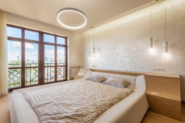 A modern bedroom with a large bed featuring light-colored bedding. Behind the bed is a textured wall with two hanging pendant lights. A circular chandelier hangs from the ceiling. The room has a large window with a view of a cityscape outside.