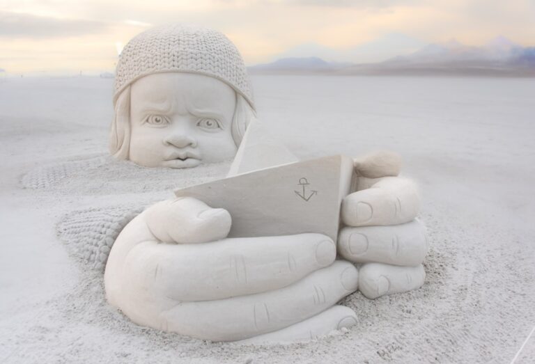 A large, detailed sand sculpture of a baby wearing a knitted hat is shown. The baby is holding a paper boat, and only its head, hands, and the boat are visible above the sand. The background is a vast, desolate landscape under a cloudy sky.