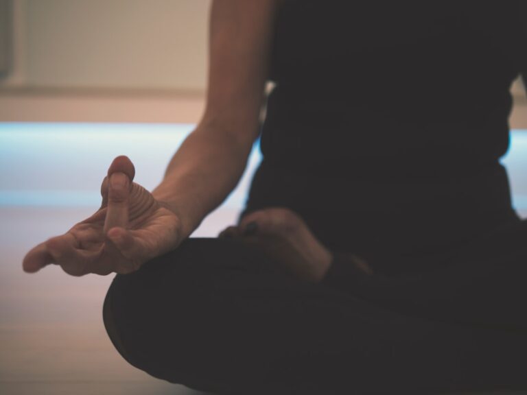 A person is sitting cross-legged on the floor, meditating. Only their torso and part of their arms and legs are visible. They are wearing dark clothing and have one hand resting on their knee with fingers positioned in a mudra gesture.