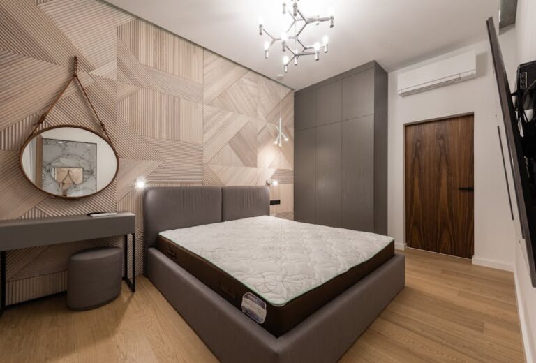 A modern bedroom featuring a large bed with a grey upholstered headboard and mattress without bedding. The room has light wooden flooring, geometric-patterned walls, a round mirror, a small desk with a stool, a large wardrobe, and a contemporary light fixture.