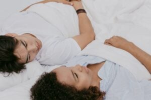 Two people lie in bed under white blankets, facing each other. One has dark hair and a white shirt, while the other has curly hair and a light blue shirt. They appear to be resting or engaged in a quiet conversation.