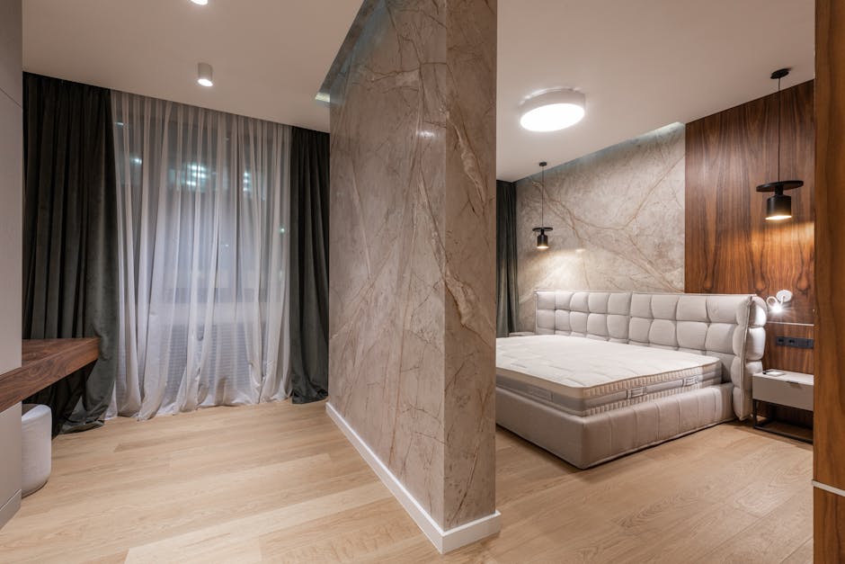 A modern bedroom with a wooden floor and a large bed with a tufted headboard. The room is divided by a marble accent wall, and features floor-to-ceiling dark curtains covering a window on the left side. Two pendant lights hang on either side of the bed.