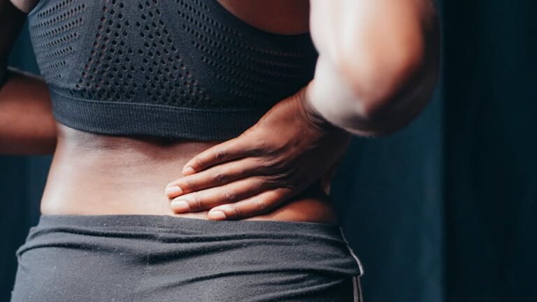 A person wearing a black sports bra and dark pants places a hand on their lower back, suggesting possible back pain or discomfort. The background is dark, highlighting the subject's posture and expression of physical relief or strain.