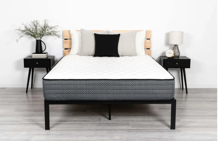 A neatly made bed showcasing the 40 Winks Studio Luxury Firm mattress and a black bed frame is flanked by two black nightstands. Each nightstand holds a lamp; the left one also has a vase with flowers, while the right one features an alarm clock. The background wall is plain white.