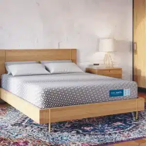 A bedroom featuring a wooden bed frame with the Diamind Gel Grid Float 10" Medium Foam Mattress neatly made with a patterned comforter. Two pillows rest on the bed, and a potted plant is nearby. Beside the bed stands a wooden nightstand with a lamp. A large closet with wooden doors is on the right, and a colorful rug covers the floor.