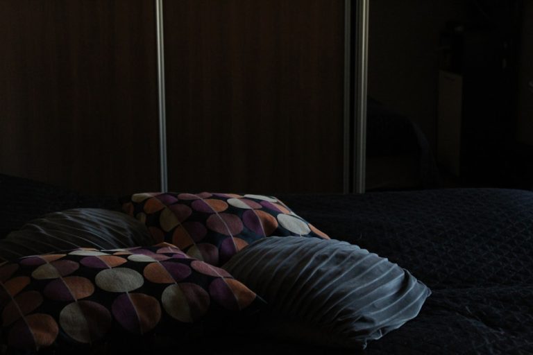 A dimly lit bedroom featuring a bed covered with a dark quilt and four pillows. Two pillows have a colorful polka dot pattern, while the other two are solid grey with a textured surface. The background shows a wooden wardrobe with vertical silver handles.