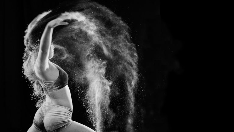 A dancer in motion, wearing a sports bra and shorts, arcs her body backward with an outstretched arm, releasing a cloud of powder that creates a dramatic, swirling effect in the dark, contrasting background.