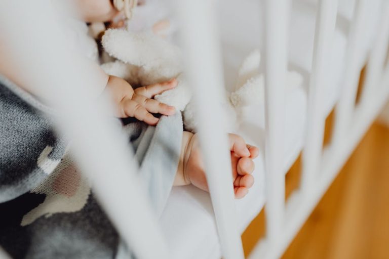 Close-up of a baby’s hand holding a soft white stuffed toy while resting in a white crib. The baby is partially covered with a blanket featuring animal designs. The image captures a peaceful and cozy moment.