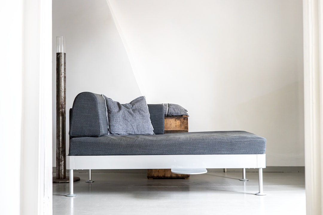 A modern minimalistic room featuring a sleek gray chaise lounge with two matching pillows. The chaise has a wooden backrest, and a tall, thin vase is situated near it. The room has white walls, a light floor, and abstracts decor elements.