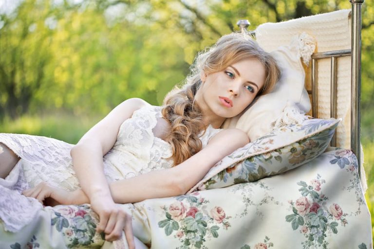 A woman with long, blonde hair in a braid lies on her side on a vintage-style bed with floral pillows and a quilted blanket. She wears a white lace dress and looks thoughtfully into the distance. The background is a sunlit outdoor setting with greenery.