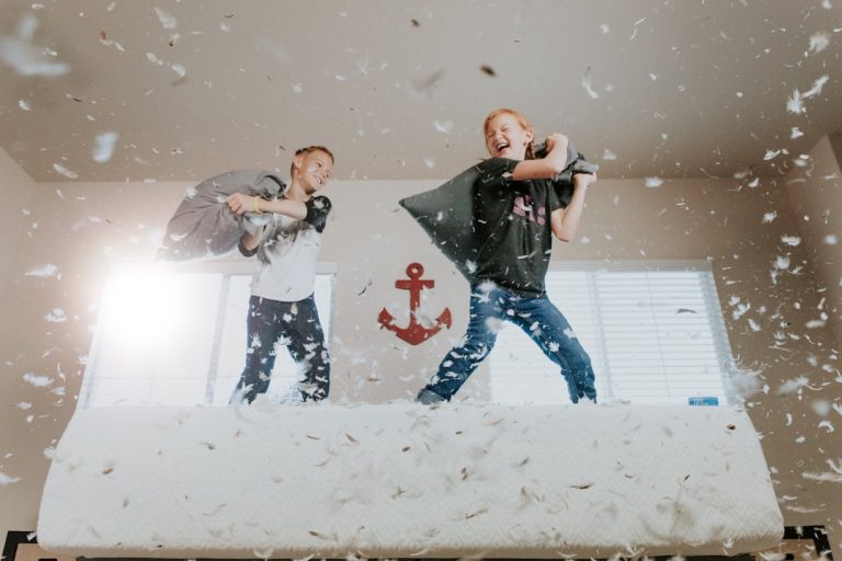 Two children are having a pillow fight on a bed in a brightly lit room. Feathers are flying everywhere, adding to the fun chaos. The room has large windows in the background, and a red anchor decoration hangs on the wall between them.