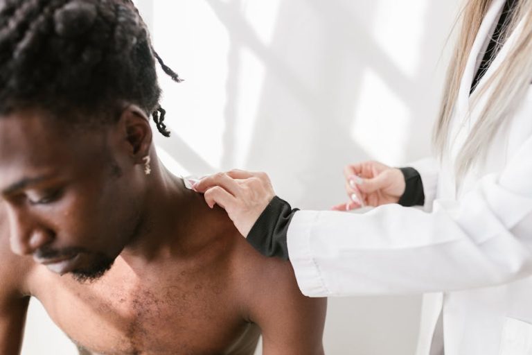 A shirtless man with braided hair sits while a person in a white coat examines his shoulder. The setting appears to be a medical office with bright, natural light.